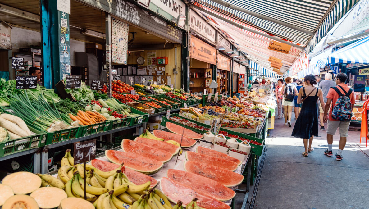 Personen auf einer Marktstraße mit Obst- und Gemüseauslagen.