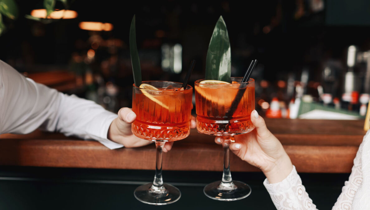 Zwei Spritz-Getränke mit Orangenscheiben in den Händen zweier Personen beim Anstoßen an einem Bartresen.