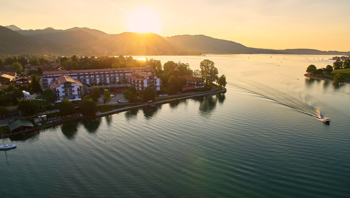 Luxuriöser Hotelkomplex an einem Seeufer, umgeben von Bäumen vor einer bewaldeten Hügellandschaft.