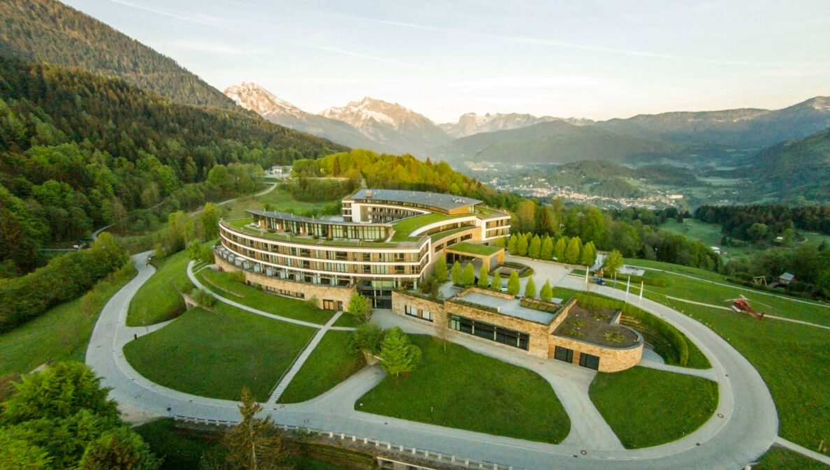 Luftaufnahme eines hufeisenförmigen Hotelkomplexes mit Rasenflächen inmitten einer hügeligen Waldlandschaft.