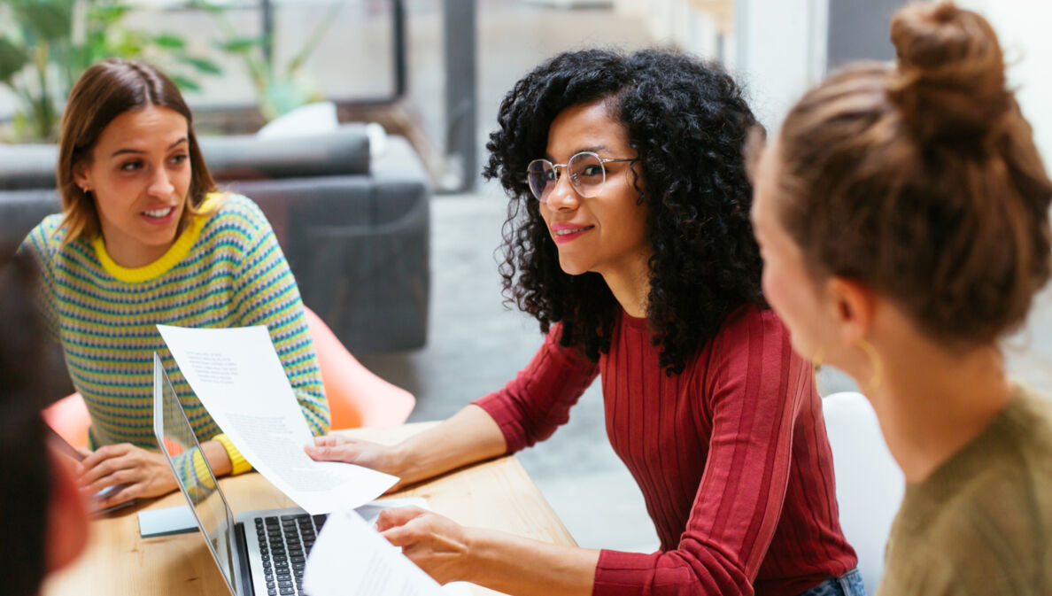 Drei junge Frauen sitzen mit Arbeitsunterlagen zusammen an einem Holztisch in einem Co-Working-Space.