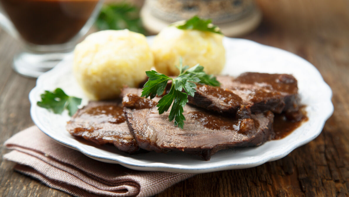 Sauerbraten mit brauner Soße an Klößen, angerichtet auf einem weißen Teller auf einem Holztisch.