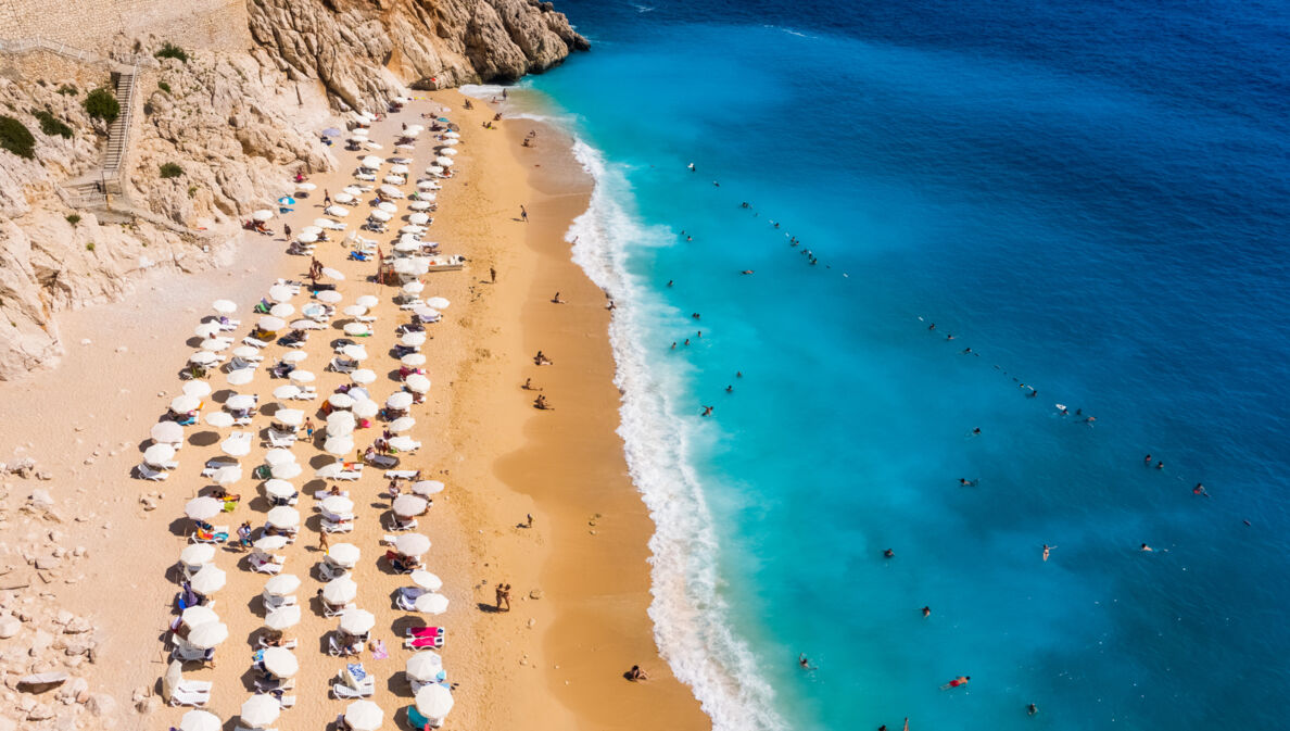 Luftaufnahme eines goldenen Sandstrandes mit Sonnenschirmen in einer Felsbucht mit Personen im türkisblauen Wasser.