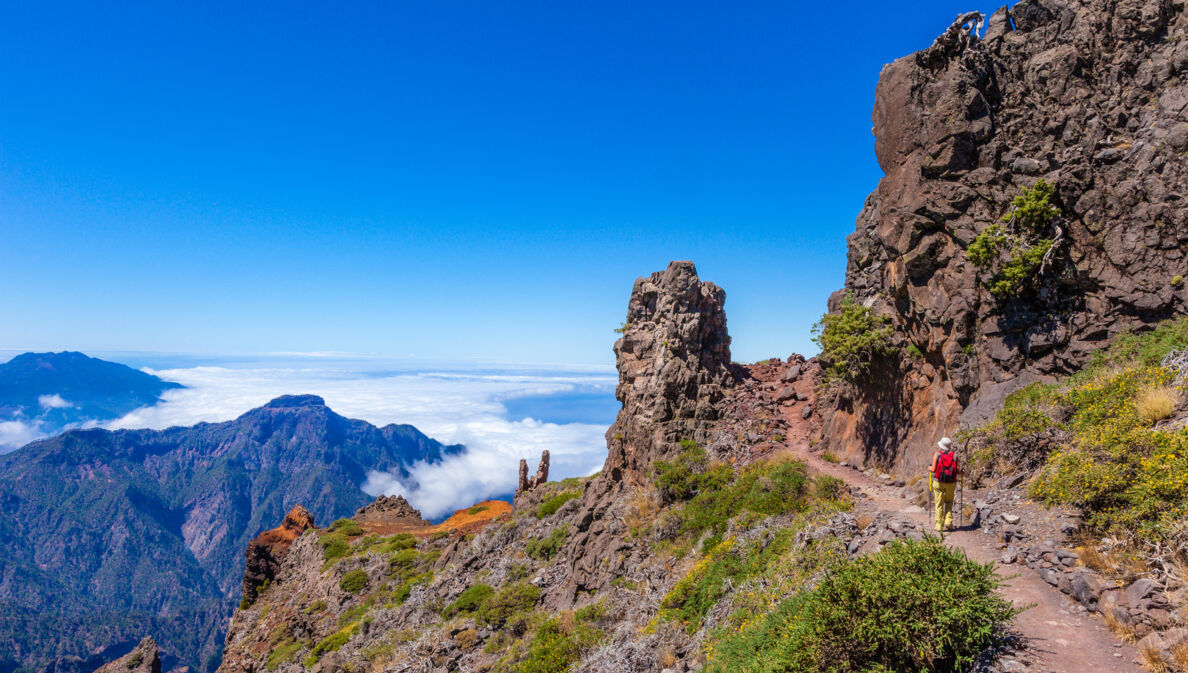 Eine Person auf einem Wanderweg auf einem Bergkamm mit rötlichen Felsformationen oberhalb der Wolkendecke unter blauem Himmel.