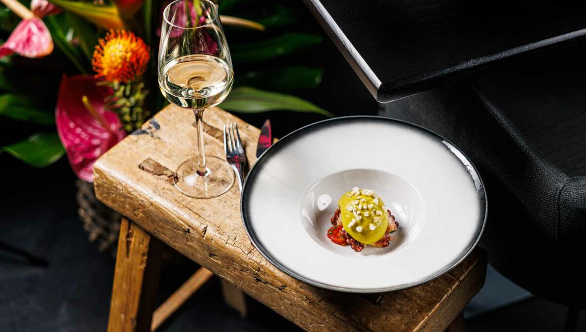 Ein weißer Teller mit einer kreativ angerichteten Vorspeise steht neben einem Glas Weißwein auf einem Holzschemel, daneben ein Blumenbouquet.