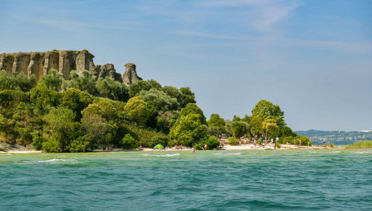 Eine antike Ruine, umgeben von üppiger Natur, an einem Sandstrand mit Personen am türkisblauen Wasser.