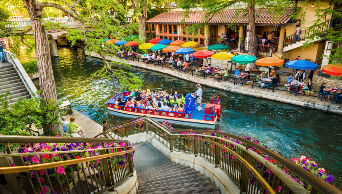 Ein Ausflugsboot fährt auf einem Wasserkanal an einem Restaurant mit Personen auf einer Terrasse mit bunten Sonnenschirmen vorbei, im Vordergrund eine mit Blumen dekorierte Treppe.