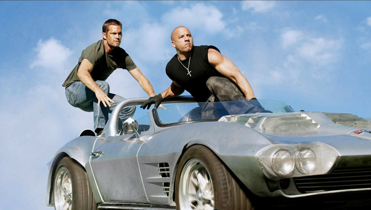 Paul Walker und Vin Diesel hocken auf einem silbernen Sportwagen ohne Verdeck.