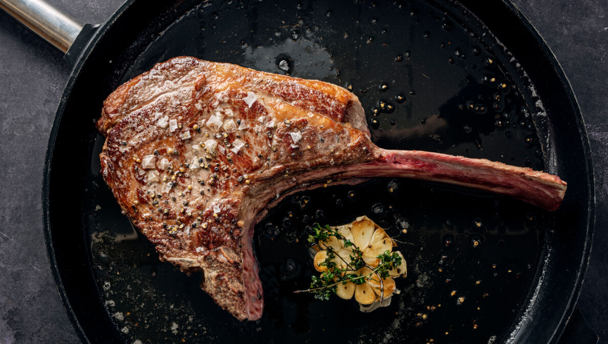 Ein scharf angebratenes Tomahawk-Steak in einer Pfanne. Daneben liegt eine aufgeschnittene Knoblauchknolle, die mit Thymian getoppt ist.