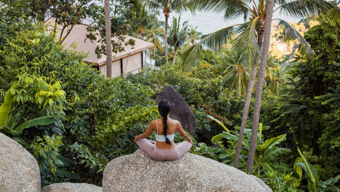 Eine Frau sitzt im Lotussitz auf einem großen Stein, vor ihr eine üppig-grüne Natur, ein Bungalow sowie der Ozean Thailands.