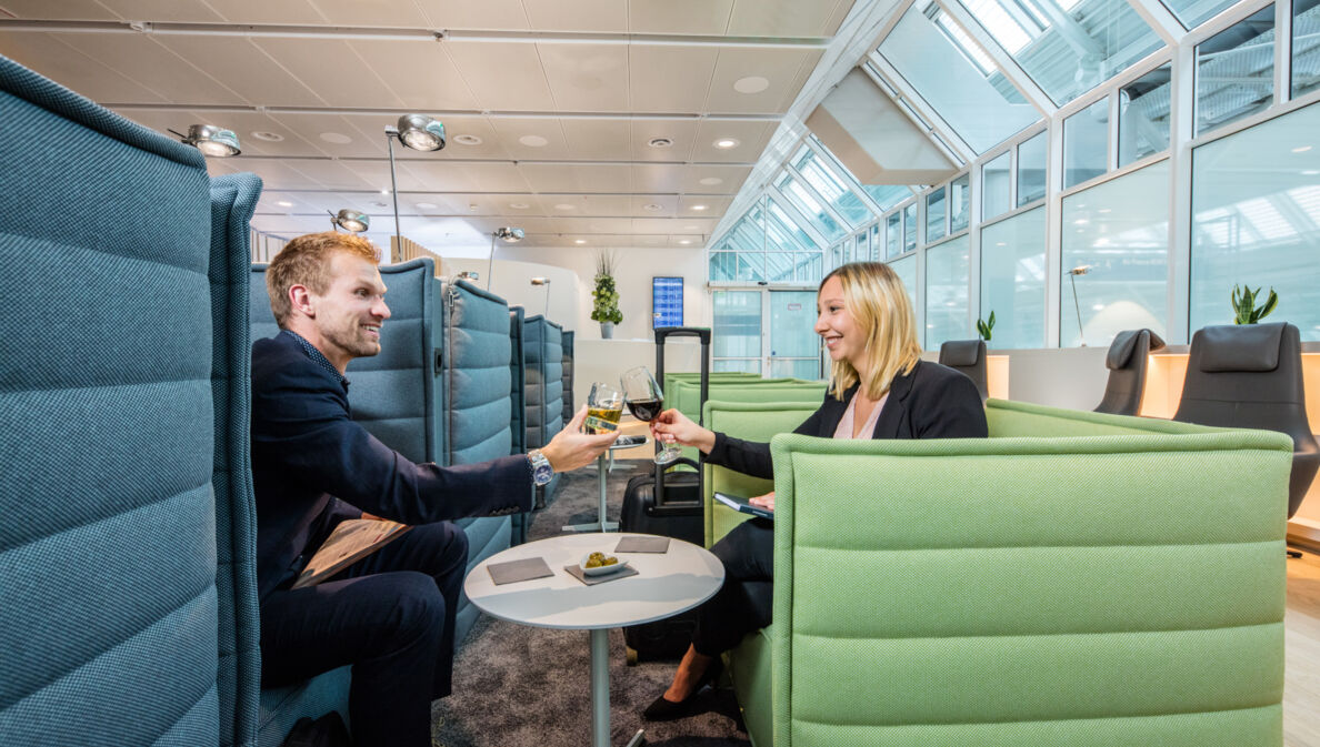 Ein Mann und eine Frau sitzen sich gegenüber auf Sesseln in einer Flughafenlounge und stoßen mit Weingläsern an.