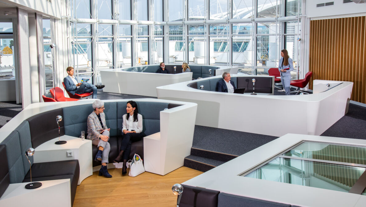 Mehrere Personen sitzen in einer modernen Sitzlandschaft einer Flughafenlounge mit Panoramafenstern.