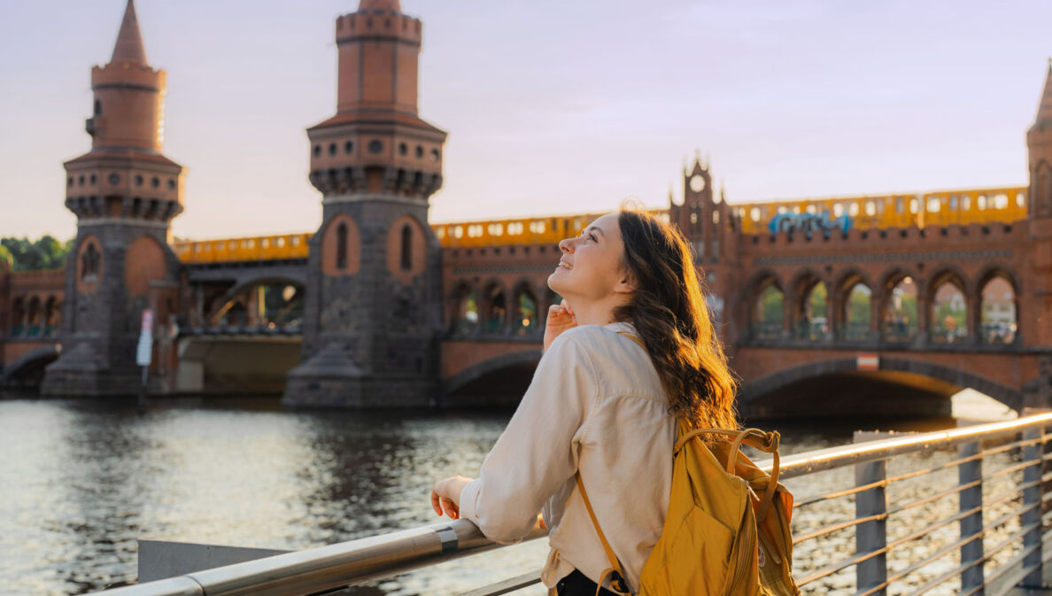 Eine junge Frau mit Rucksack steht am Wasser vor einer Brücke mit Türmen, auf der eine gelbe Straßenbahn fährt.