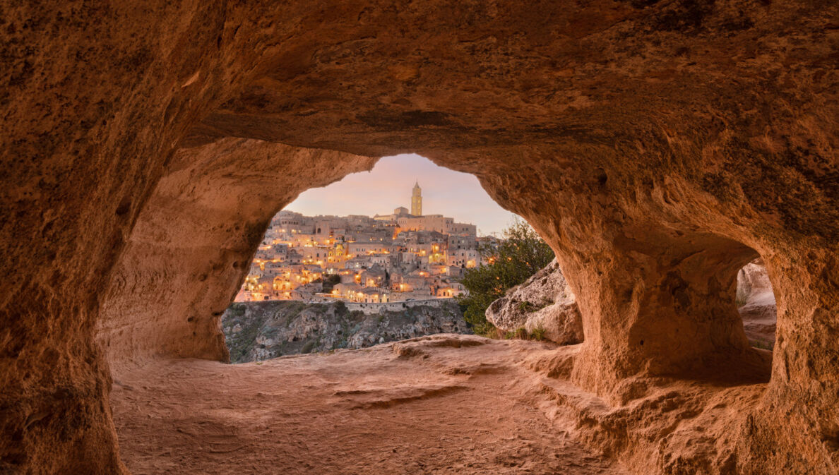 Blick auf die erleuchtete Stadt Matera aus einer Steingrotte im Vordergrund bei Dämmerung.