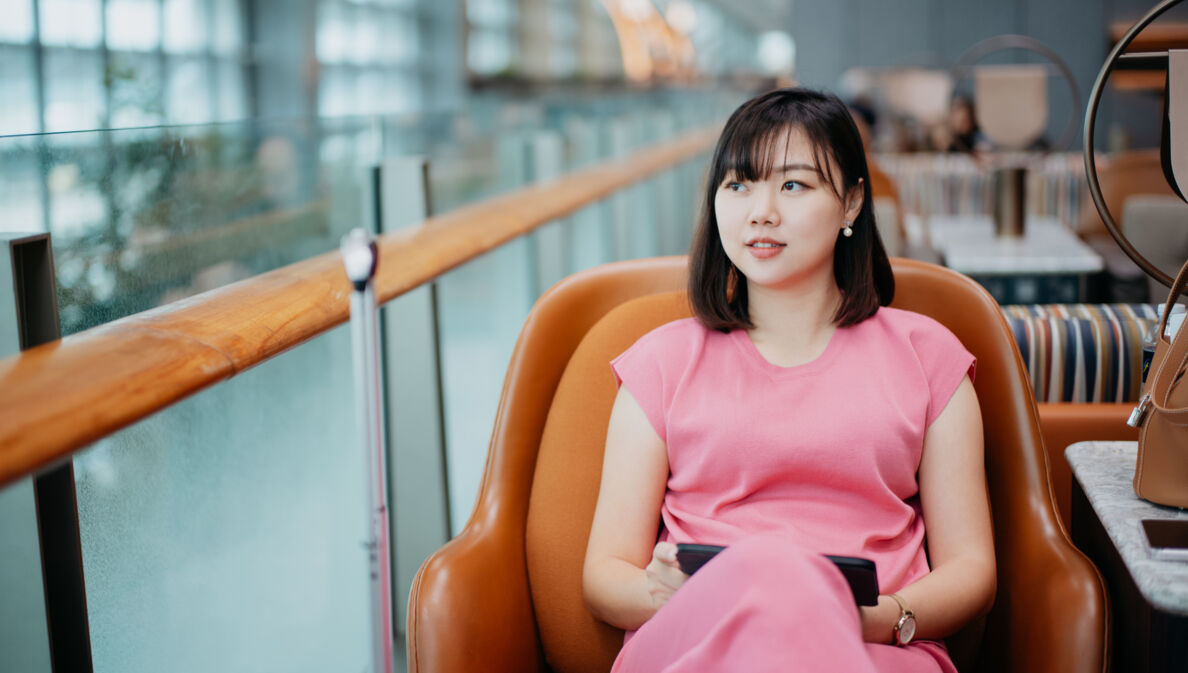 Eine asiatische Frau in einem pinken Kleid sitzt in einem braunen Ledersessel auf einer Galerie einer Flughafenlounge.