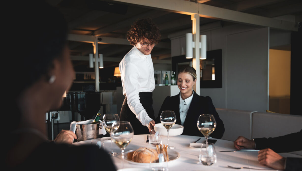 Eine lächelnde Frau wird in einem Restaurant von einem Kellner bedient, der einen Teller vor ihr auf den weiß eingedeckten Tisch stellt.