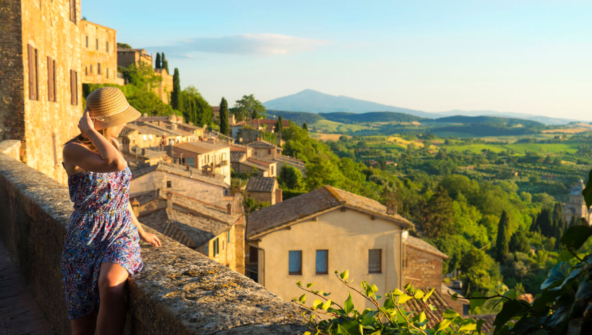 Eine Frau im Sommerkleid blickt von einer Mauer in einem italienischen Dorf hinunter auf eine grüne Hügellandschaft bei warmem Sonnenlicht.