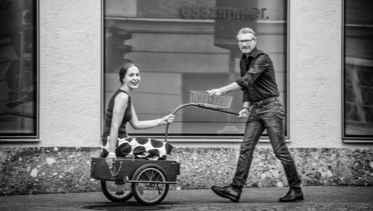 Ein Mann zieht eine lächelnde Frau in einem Fahrradanhänger auf einem Gehweg vor einer Fensterfront.