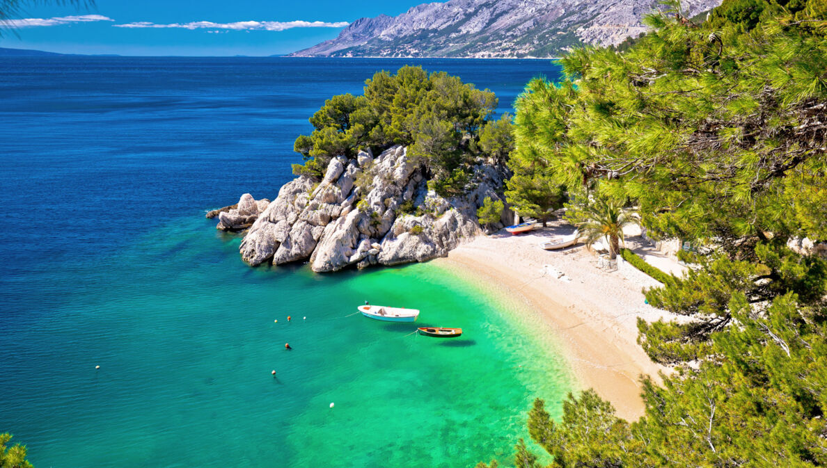 Kleine Sandbucht mit Fischerbooten im türkisblauen Meer, umgeben von Felsen und Kiefernwald.