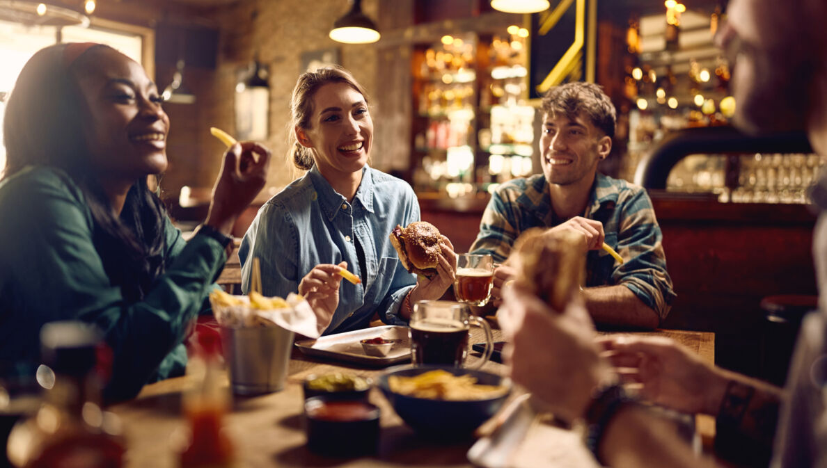 Vier junge Menschen an einem Holztisch essen Fastfood in einem rustikalen Restaurant.