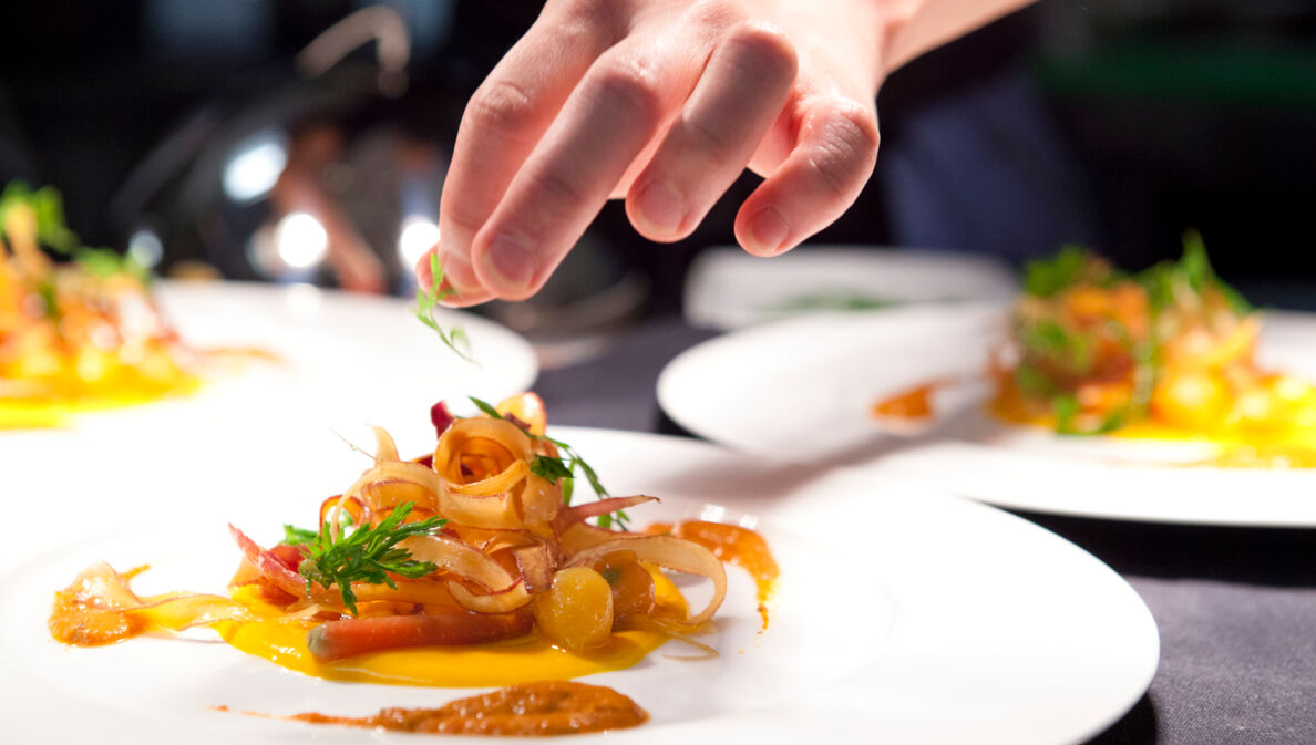 Eine Hand garniert ein Gericht auf einem weißen Teller in einer Restaurantküche.