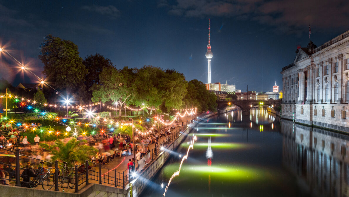 Personen in einer Strandbar am Spreeufer an der Berliner Museumsinsel bei Nacht, im Hintergrund ein beleuchteter Fernsehturm.