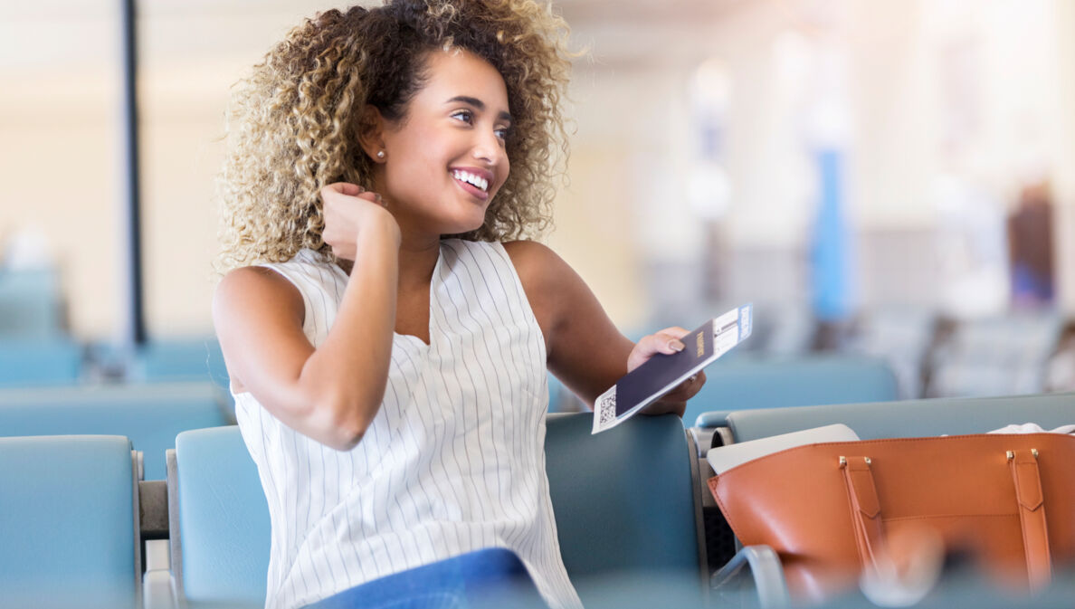 Eine junge, lächelnde Frau sitz mit Reisepass in der Hand in einer Flughafenlounge.