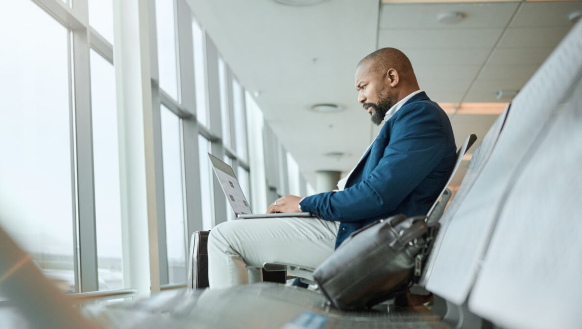 Ein Mann, der am Flughafen am Fenster sitzt und einen aufgeklappten Laptop auf dem Schoß hat.