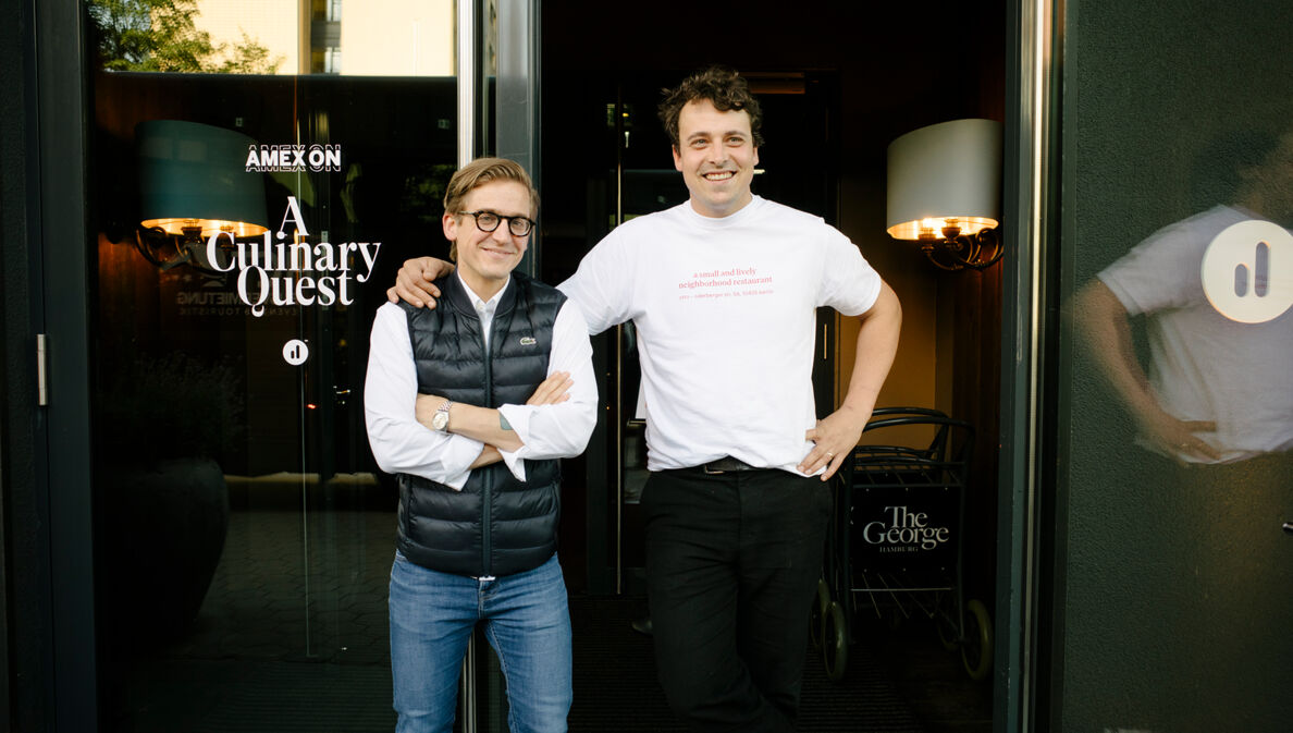 Zwei Männer posieren vor einer Glastür mit der Aufschrift „AMEX ON A Culinary Quest“ am Eingang eines Hotels.