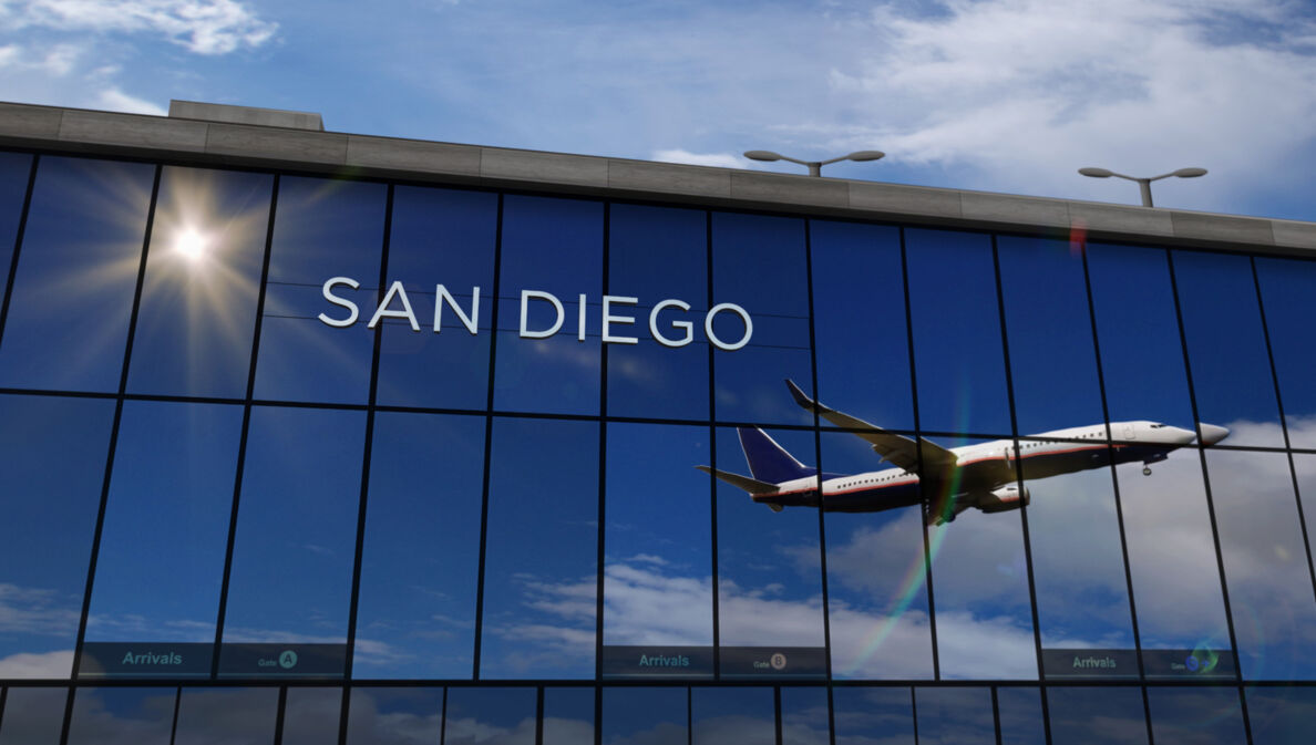 Flugzeug beim Abflug, das sich in den verspiegelten Fenstern einer Flughafenhalle spiegelt, auf denen die Aufschrift „San Diego“ zu lesen ist.