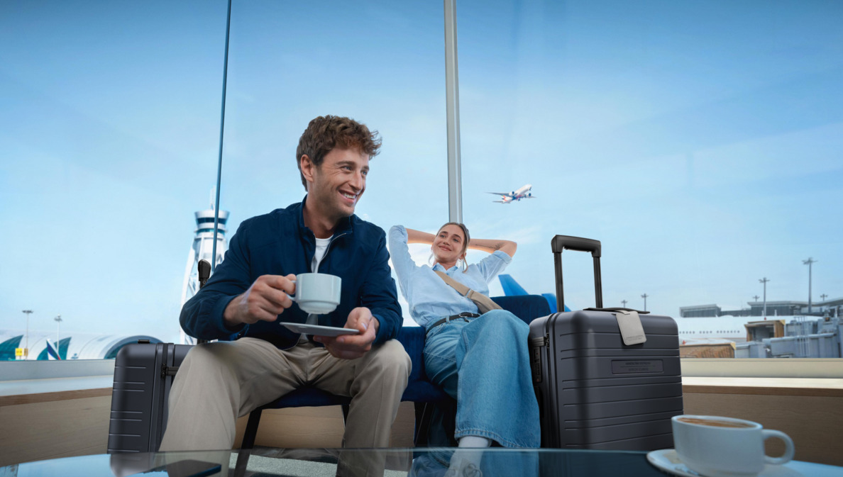 Zwei Personen sitzen nebeneinander in einer Flughafen-Lounge, neben sich jeweils einen Rollkoffer der Marke HORIZN, und trinken einen Kaffee.