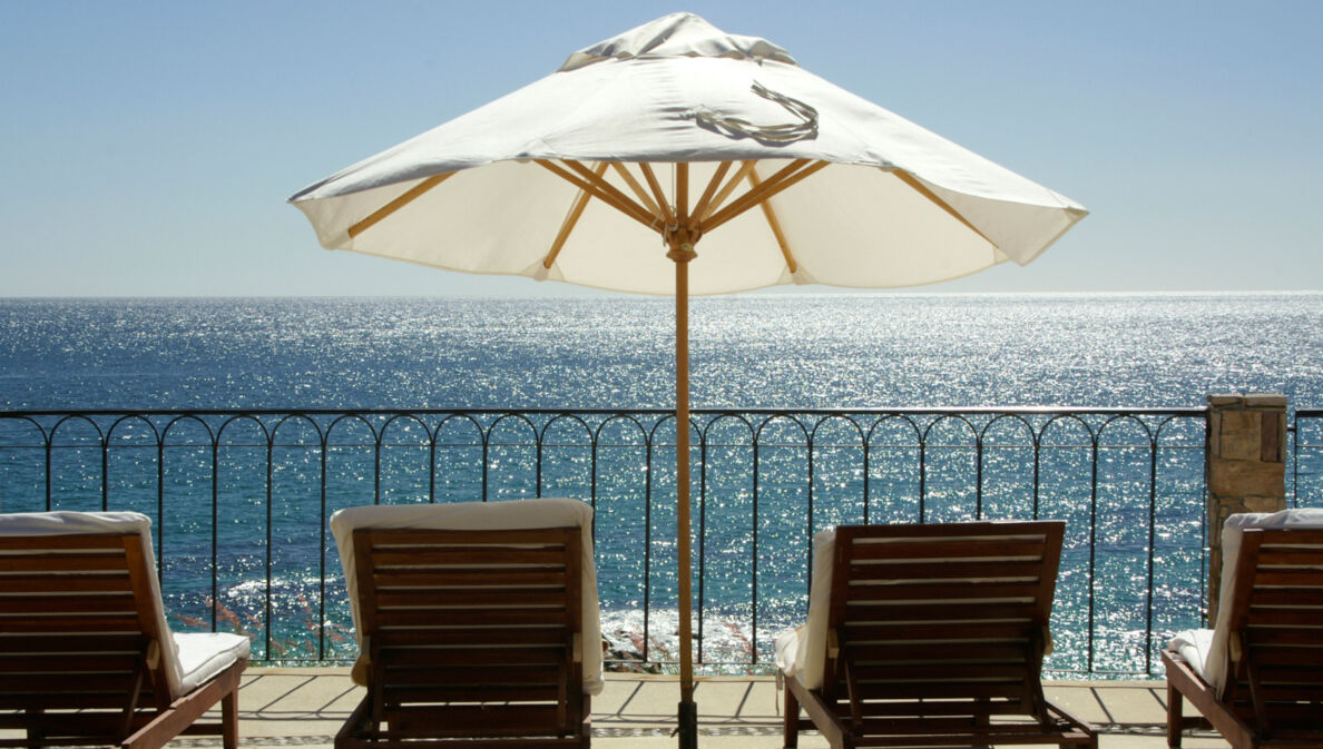 Terrasse und Geländer mit Blick aufs Meer, auf der mehrere Liegestühle und ein weißer Sonnenschirm stehen.