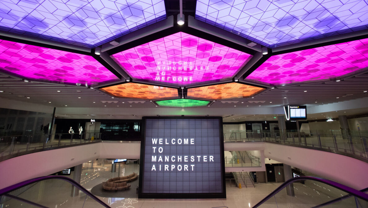 Menschenleere Flughafenhalle mit einem Bildschirm im Mittelpunkt, auf dem „Welcome to Manchester Airport“ steht und bunt erleuchteten LED-panels an der Decke.