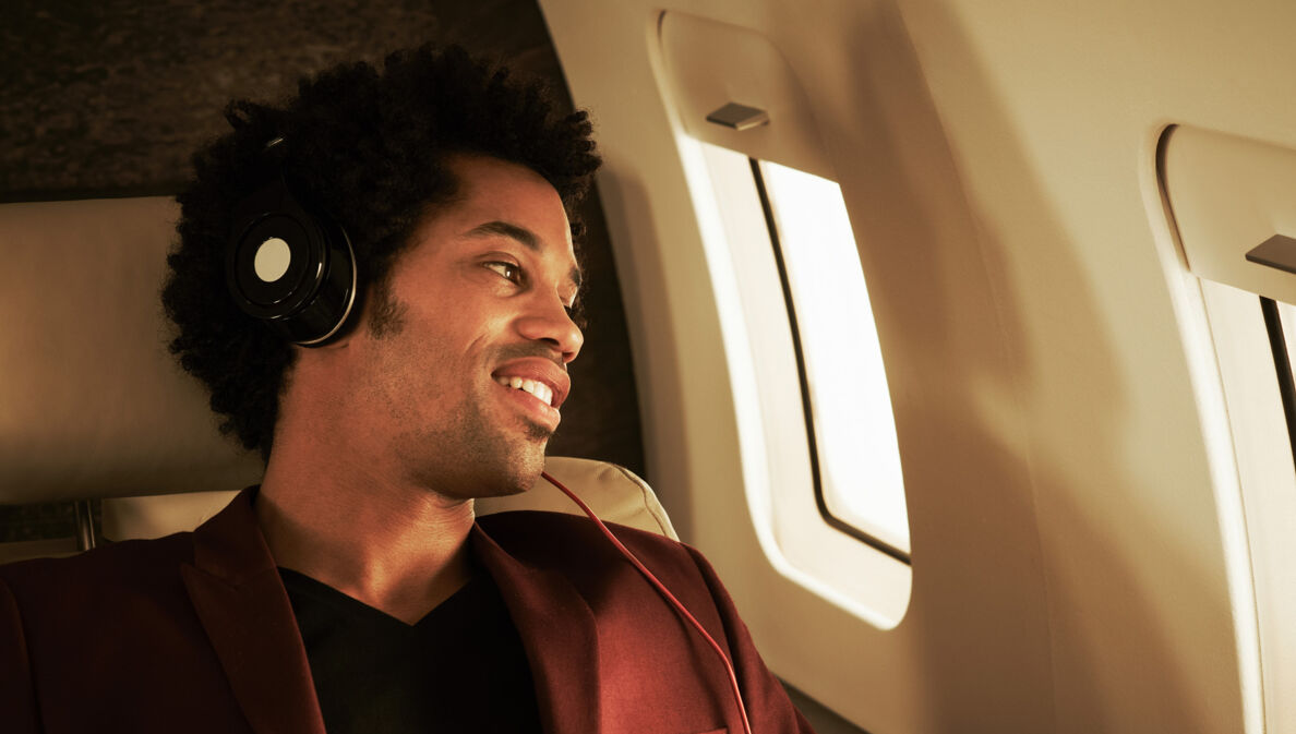 Ein junger dunkelhäutiger Mann mit Kopfhörern schaut lächelnd aus dem Fenster in einem Flugzeug.