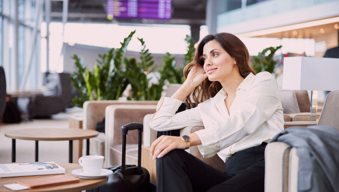 Eine Frau in Business-Kleidung sitzt in einem hellen Lounge-Sessel. Neben ihr steht ein Koffer, auf dem Tisch vor ihr befinden sich Reisedokumente und eine Tasse mit Untersetzer.