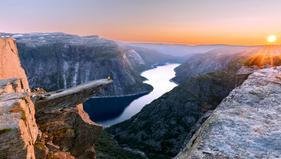 Fjordlandschaft mit Person auf einer Felsklippe bei Sonnenuntergang.