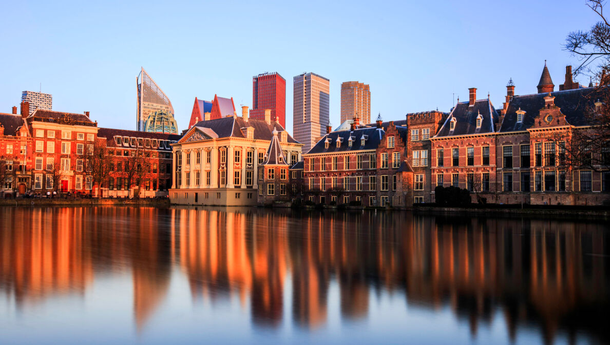 Skyline von Den Haag bei Sonnenuntergang, die Gebäude spiegeln sich im Wasser.