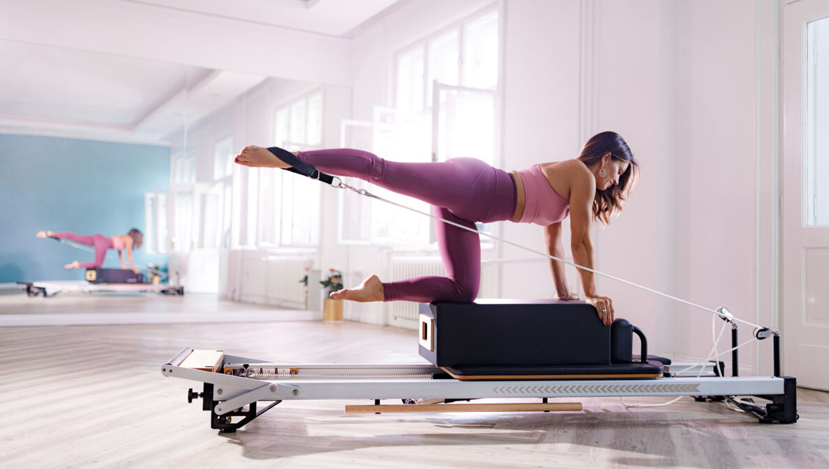 Eine Frau in pinker Sportkleidung kniet auf einer Box auf einem Pilates Reformer in einem hellen Raum.