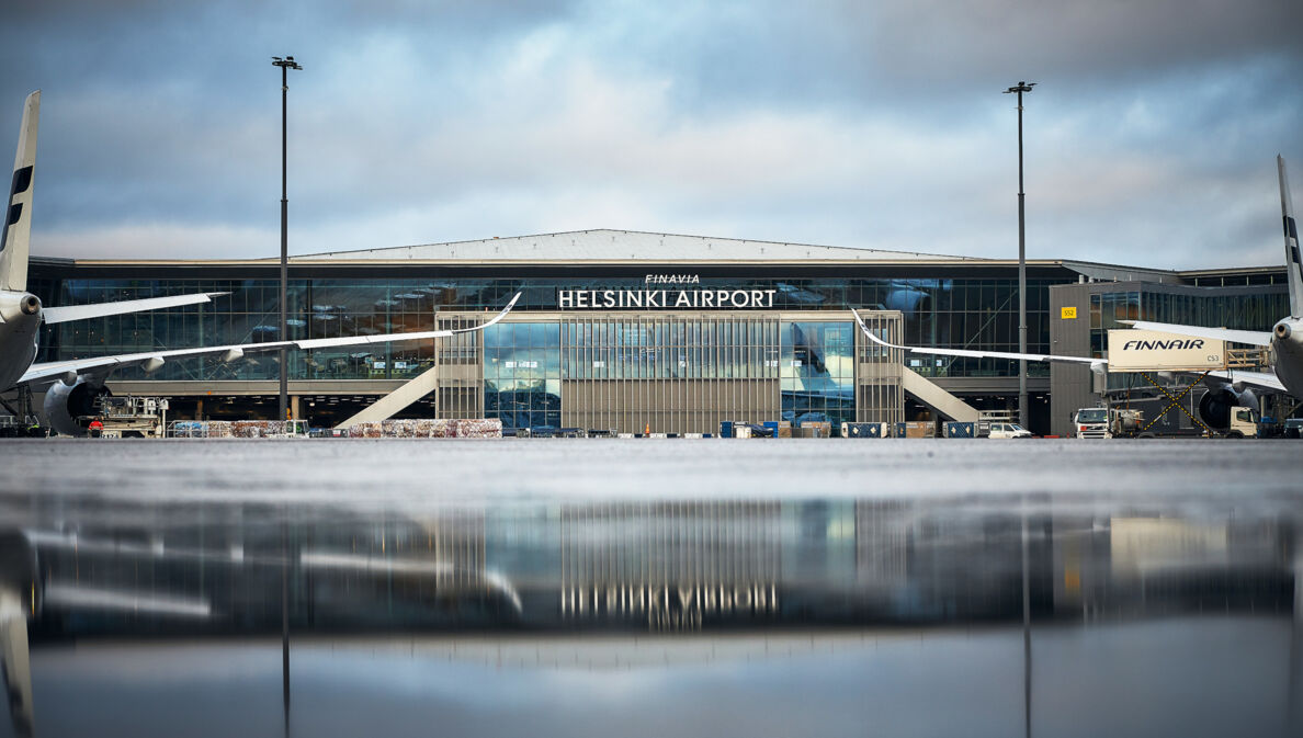 Helsinki Flughafenterminal mit Flugzeugen, im Vordergrund Spiegelung des Gebäudes auf nassem Rollfeld.