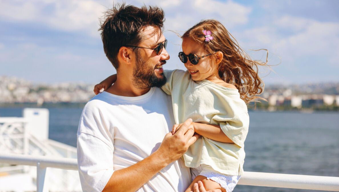 Ein lächelnder Mann mit Sonnbrille mit einem fröhlichen Mädchen mit Sonnenbrille auf dem Arm auf dem Außendeck eines Schiffes.