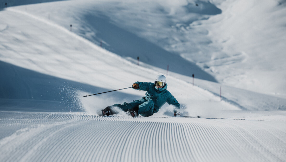 Ein Skifahrer mit Helm im türkisen Schneeanzug bei der Abfahrt auf einer Piste.