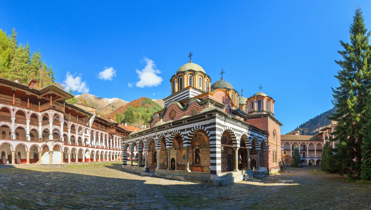 Orthodoxe Klosteranlage mit farbenfrohen Gebäuden im Gebirge unter blauem Himmel.