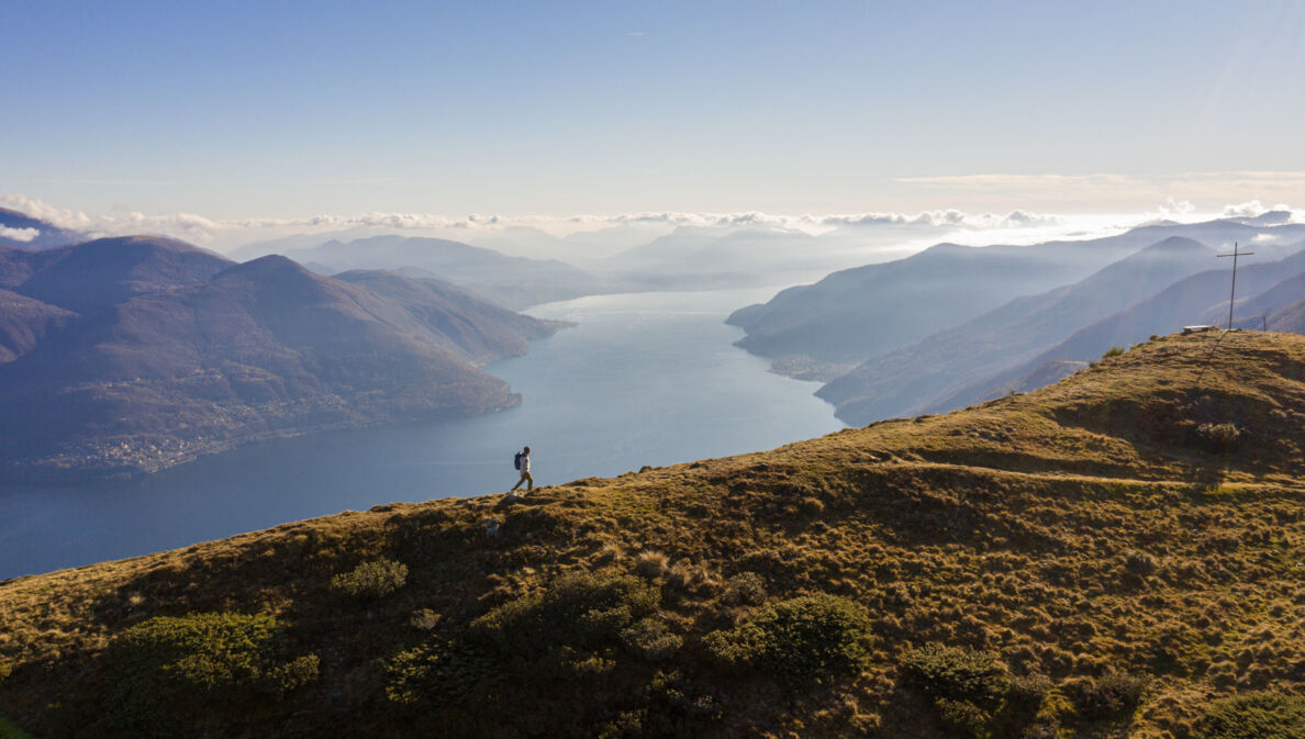 Panoramaaufnahme von einem Mann mit Rucksack auf einem Berg und dem Lago Maggiore im Hintergrund.