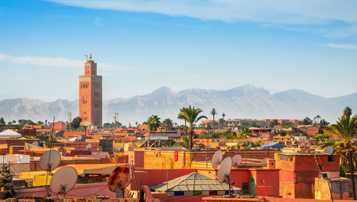 Stadtpanorama von Marrakesch mit Minarett vor Gebirgskette im Hintergrund unter blauem Himmel.