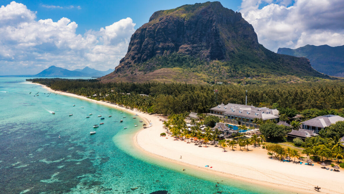 Küstenpanorama mit Hotelresort an einem palmengesäumten, weißen Sandstrand an türkisblauem Meer, im Hintergrund Berge.