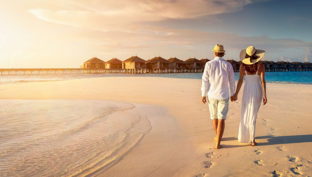 Rückansicht eines händchenhaltenden Paares in heller Sommerkleidung und mit Strohhüten an einem Strand vor einem Holzstegmit Wasserbungalows.