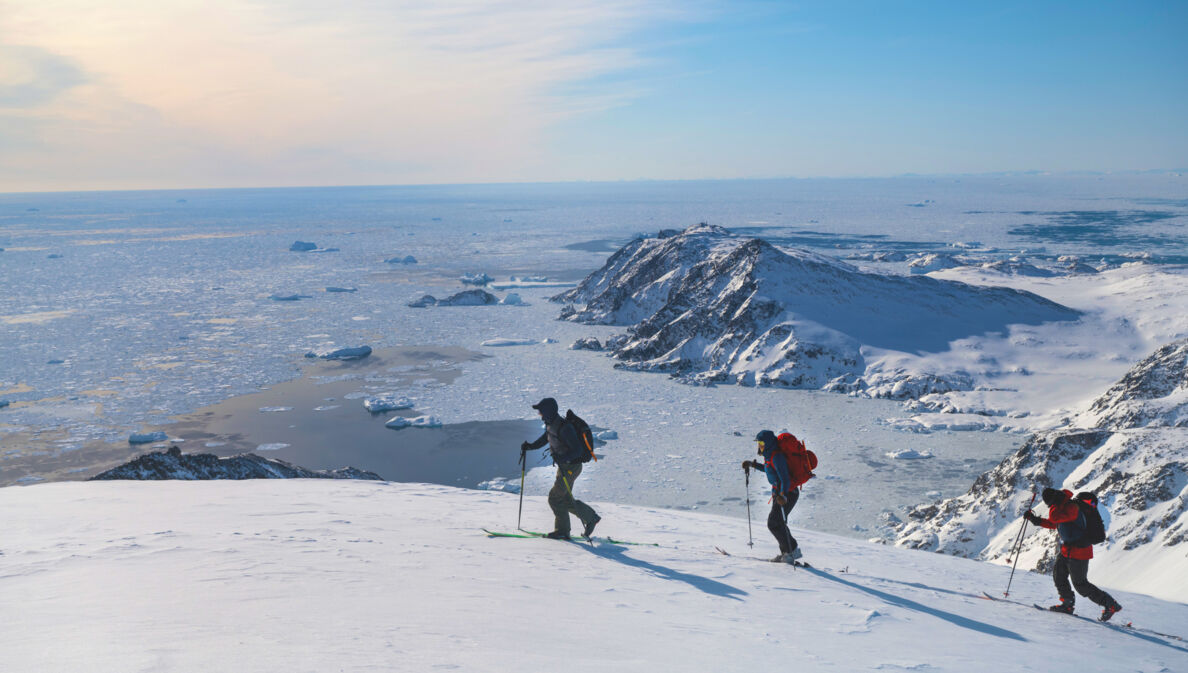 Drei Personen auf Skiern laufen einen schneebedeckten Bergkamm vor einer Gletscherlandschaft entlang.