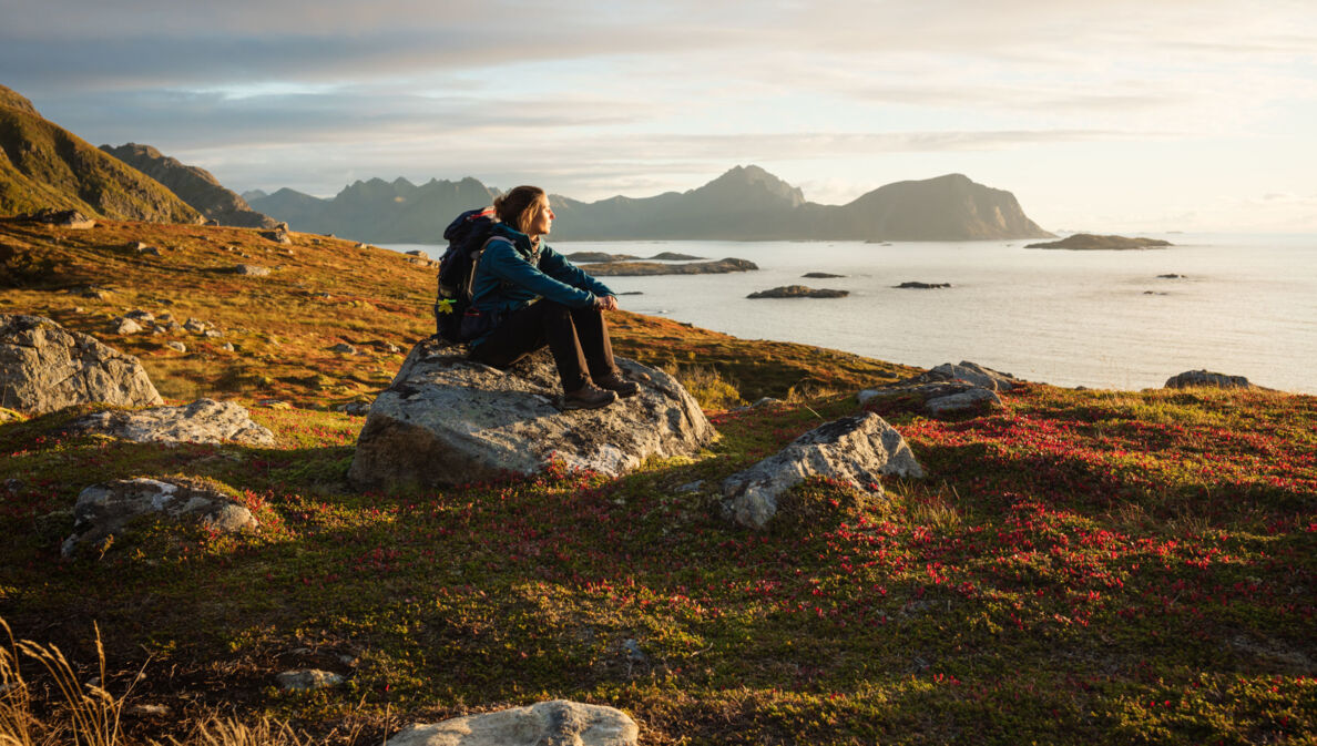 Eine Frau mit Wanderausrüstung, die bei Sonnenuntergang auf einem großen Stein sitzt und aufs Wasser blickt, im Hintergrund Berge.