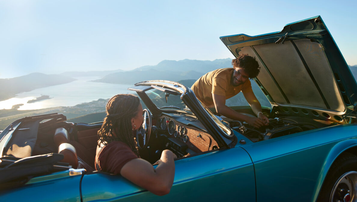 Ein Paar in einem liegengebliebenen Cabriolet an einer Meeresküste bei Sonnenschein, er kontrolliert den Motor, sie sitzt auf dem Beifahrersitz.