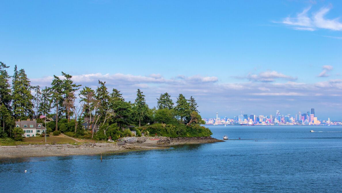 Vereinzelte Häuser auf einem schmalen Inselabschnitt, umgeben von Wasser vor Seattles Skyline im Hintergrund.